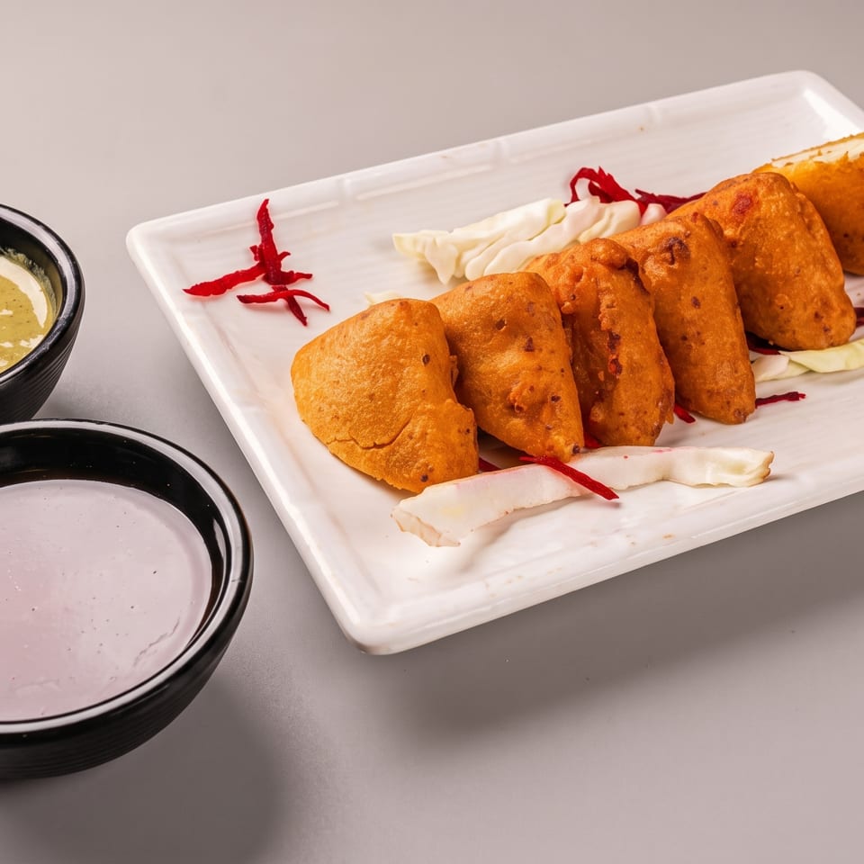 Fried paneer pakora on a white plate with two bowls of dipping sauces.